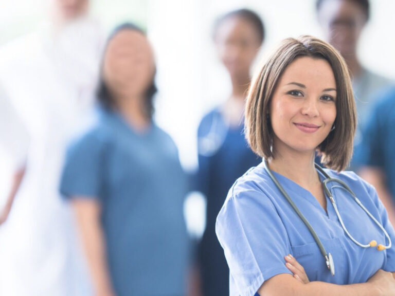 Smiling nurse with stethoscope, team blurred background.