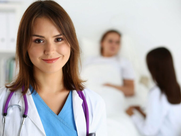 Doctor smiling, patient in background, medical setting.